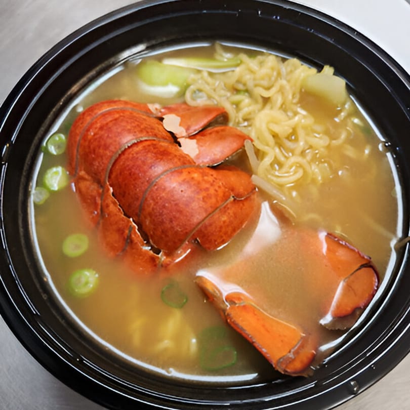 A black bowl of Korean ramen with a large lobster tail, noodles, green onions, and bok choy.