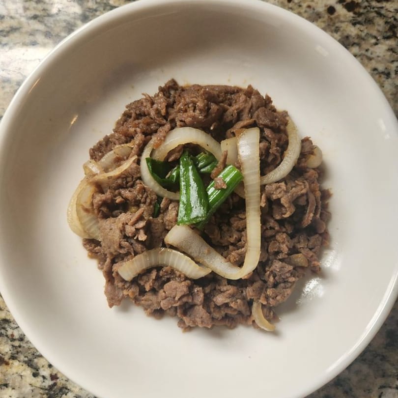 A white bowl filled with Korean bulgogi, topped with sliced onions and green onions.